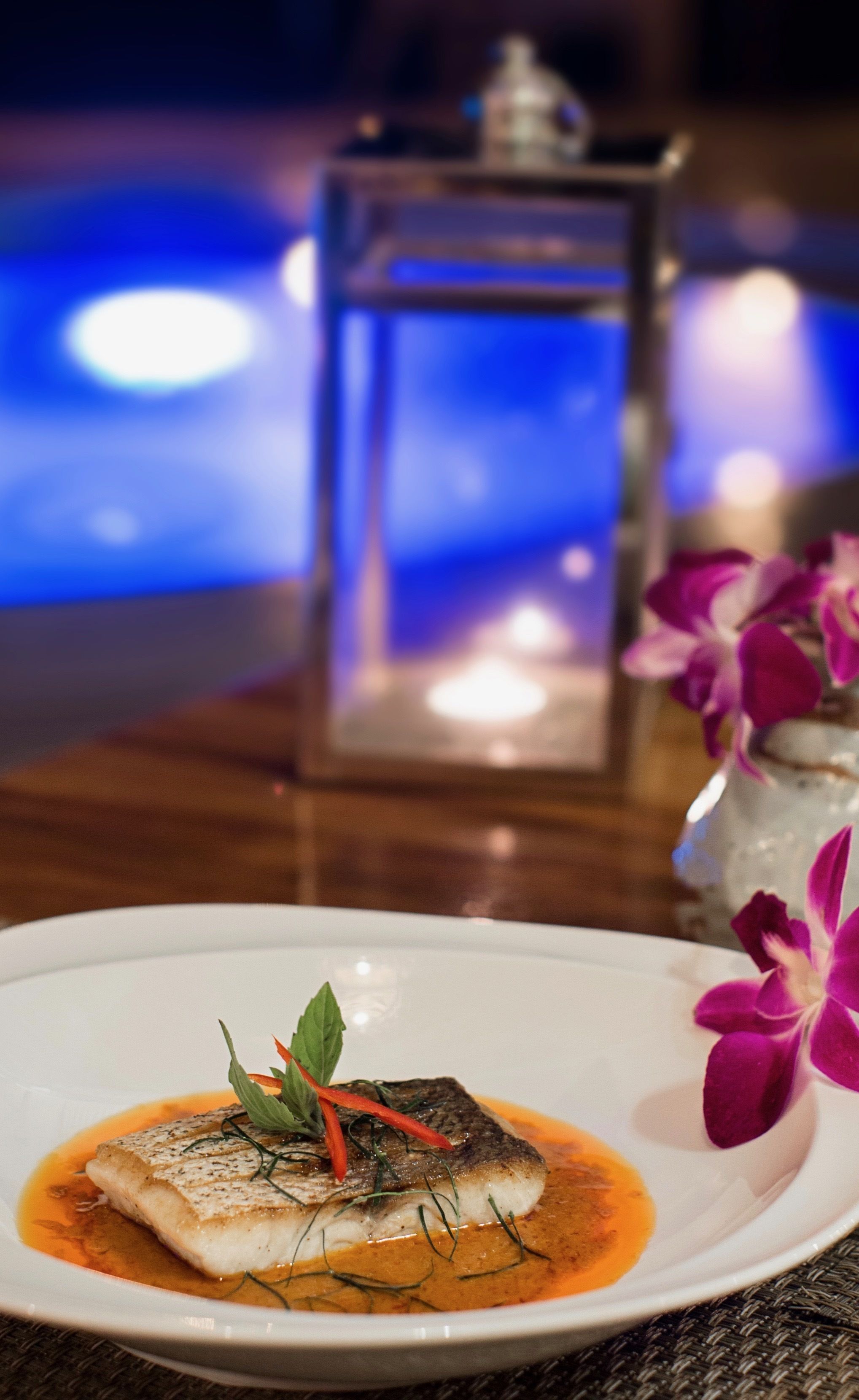 Un plato de pescado con salsa y decoración, una vela y una orquídea al fondo.