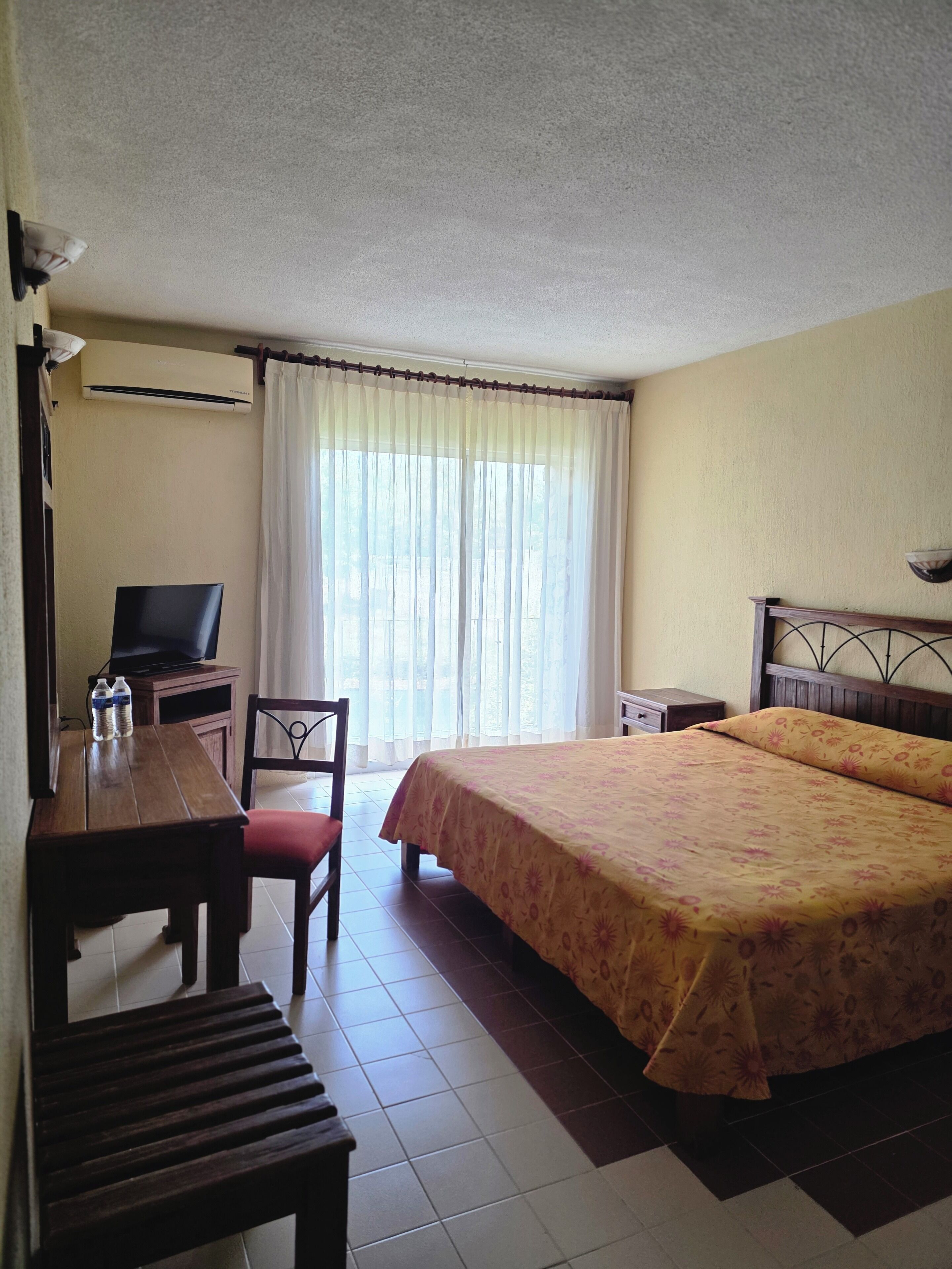 A hotel room with a bed, a desk with a television, a chair, and a window with curtains.
