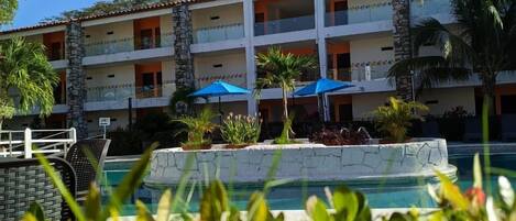 A hotel with a pool, surrounded by tropical plants and umbrellas.