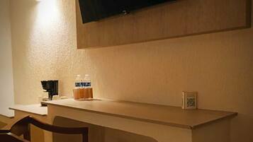 Une chambre dotée d’un téléviseur à écran plat fixé au mur, d’un bureau en bois avec une chaise, et d’une petite table sur laquelle se trouvent une bouteille et une boîte.