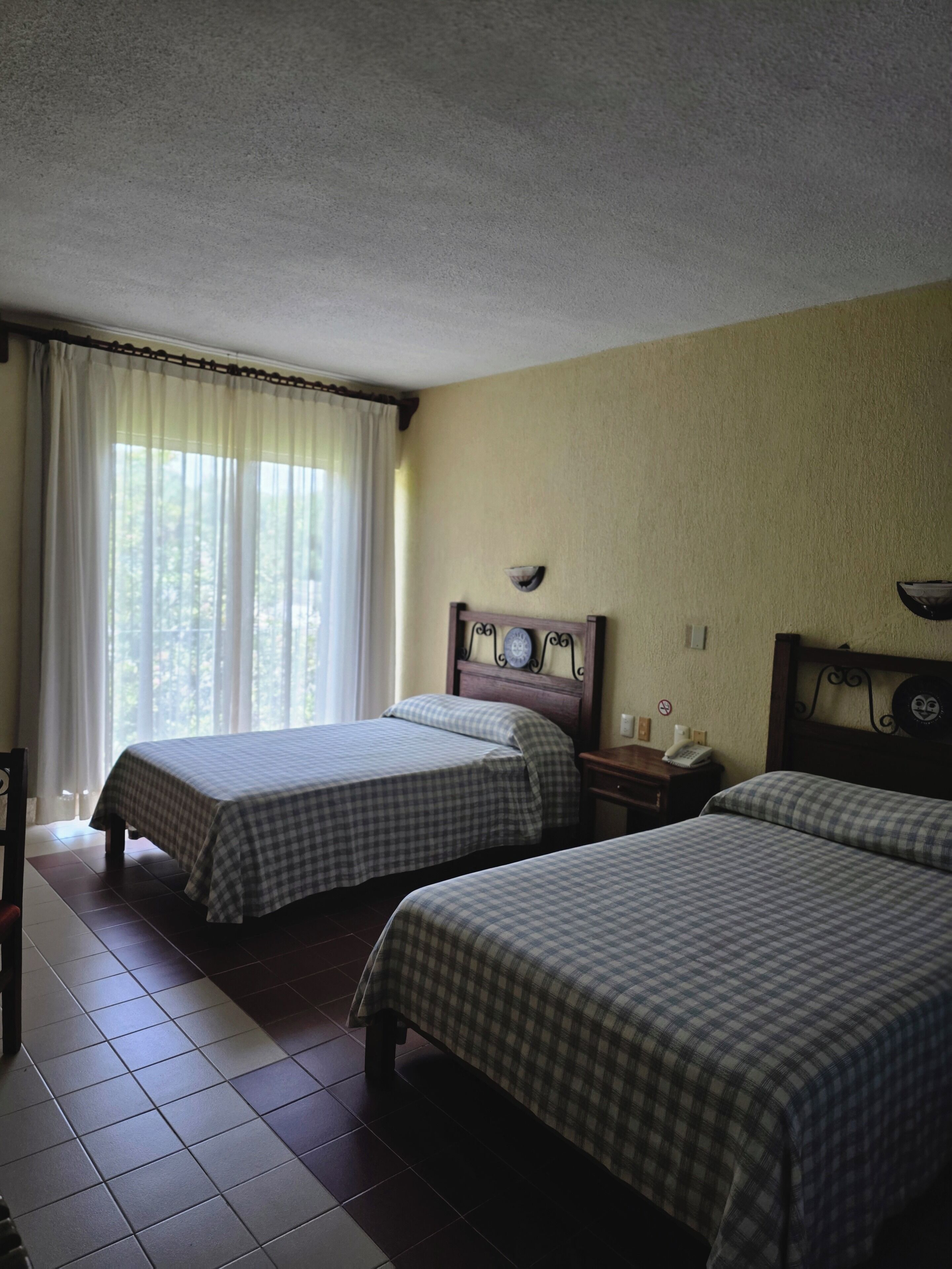 A room with two beds, a wooden headboard, a bedside table with a lamp, and a window with curtains.