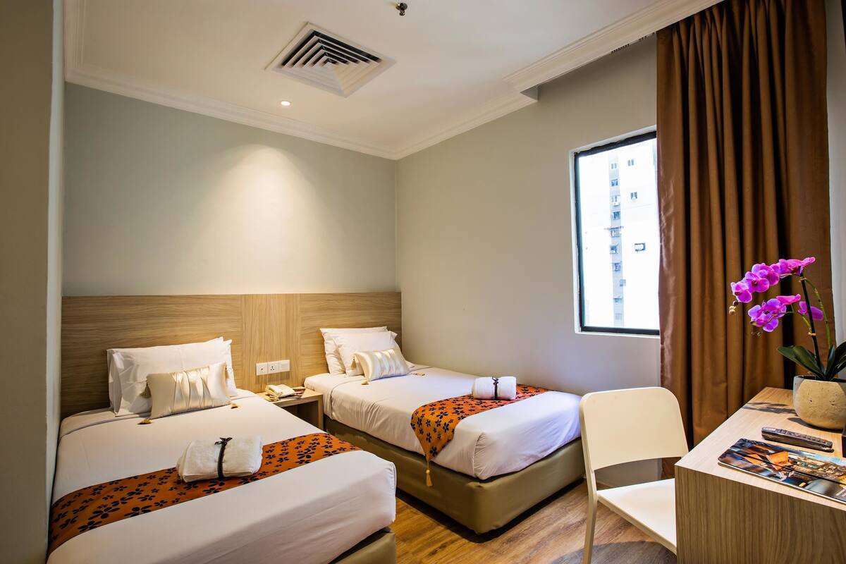 A hotel room with two beds, a desk, a chair, a window with curtains, and a potted plant.