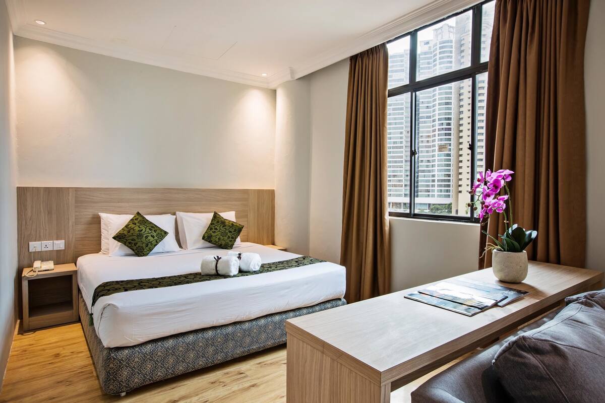 A hotel room with a neatly made bed, a bedside table with a vase of flowers, and a view of the city through large windows.