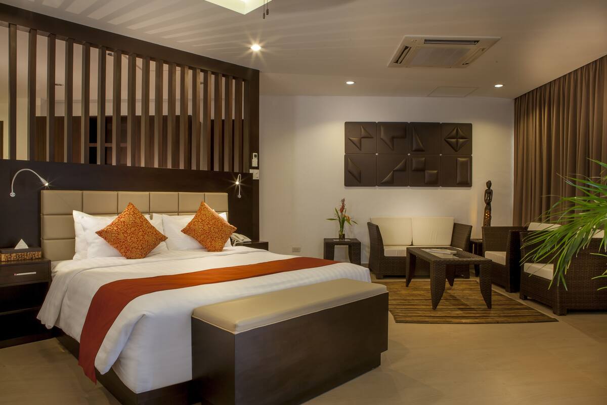 A modern hotel room with a large bed, a seating area with a sofa and armchairs, a small table, and a decorative wall piece.