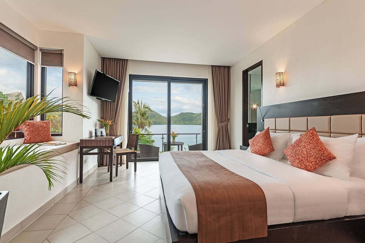 A hotel room with a large bed, a desk with a chair, a TV, and a balcony with a view of the hills.