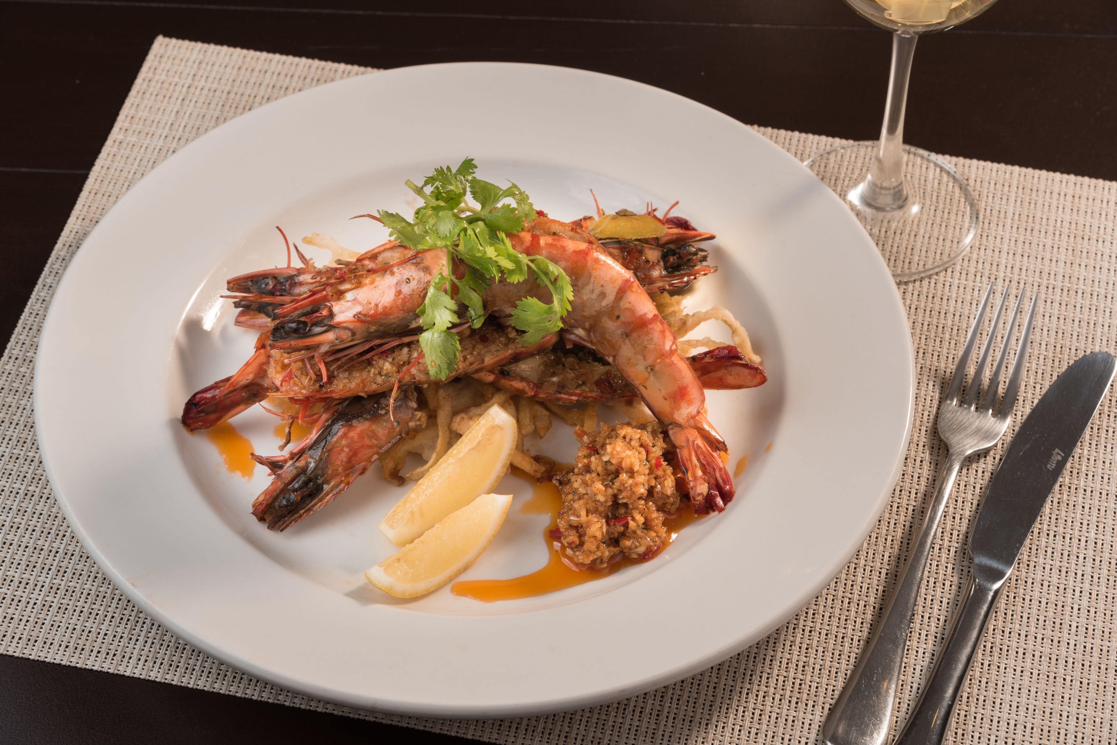 A plate of cooked shrimp with lemon wedges and a garnish, placed on a table with a fork and knife beside it.
