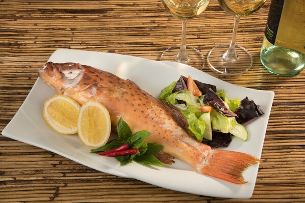 A plate of cooked fish with lemon slices and herbs, accompanied by a salad and two glasses of white wine on a wooden table. - Busuanga Bay Lodge (Busuanga)