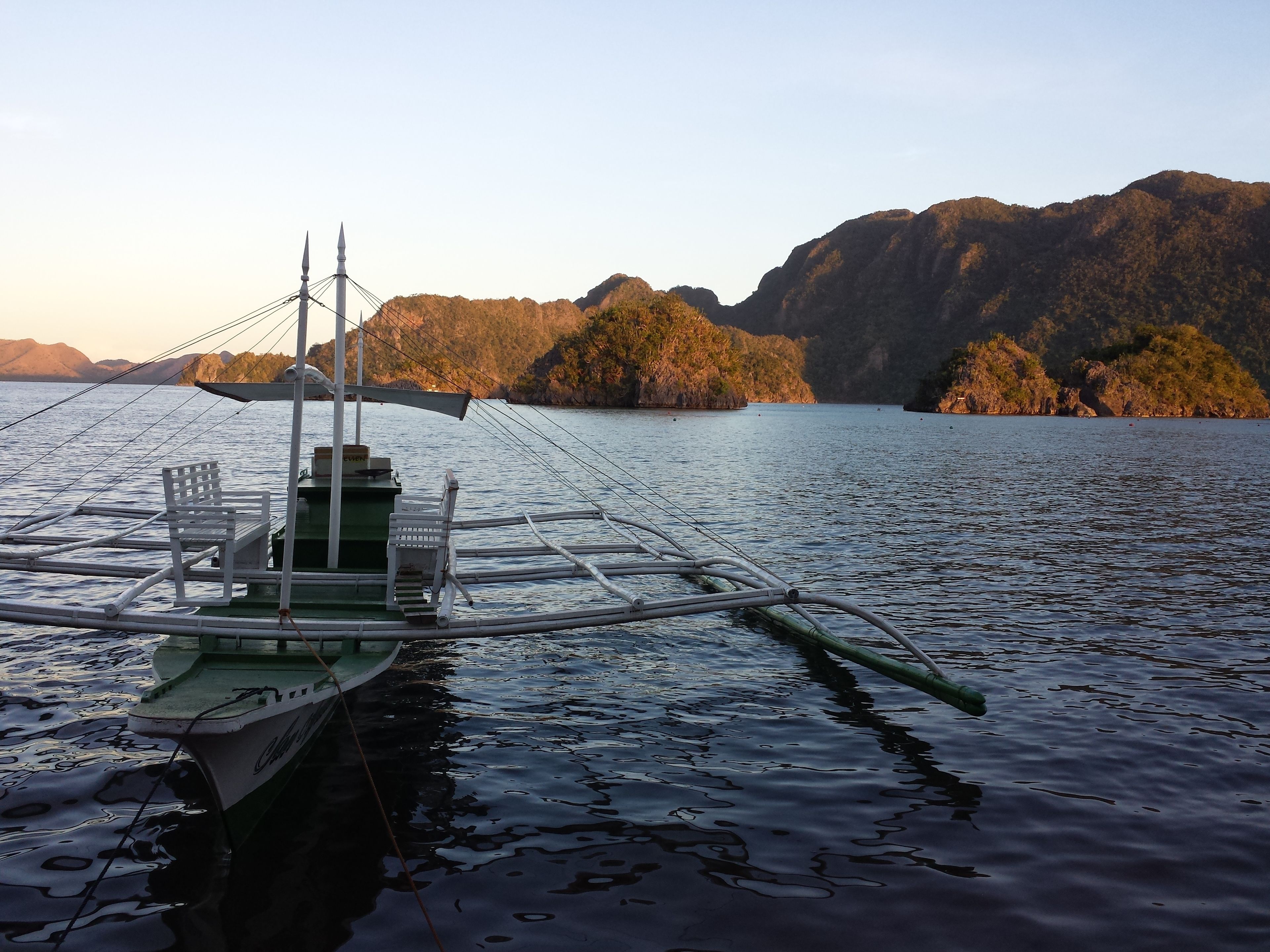 A boat with a green hull and white deck, anchored in calm waters with a mountainous landscape in the background.