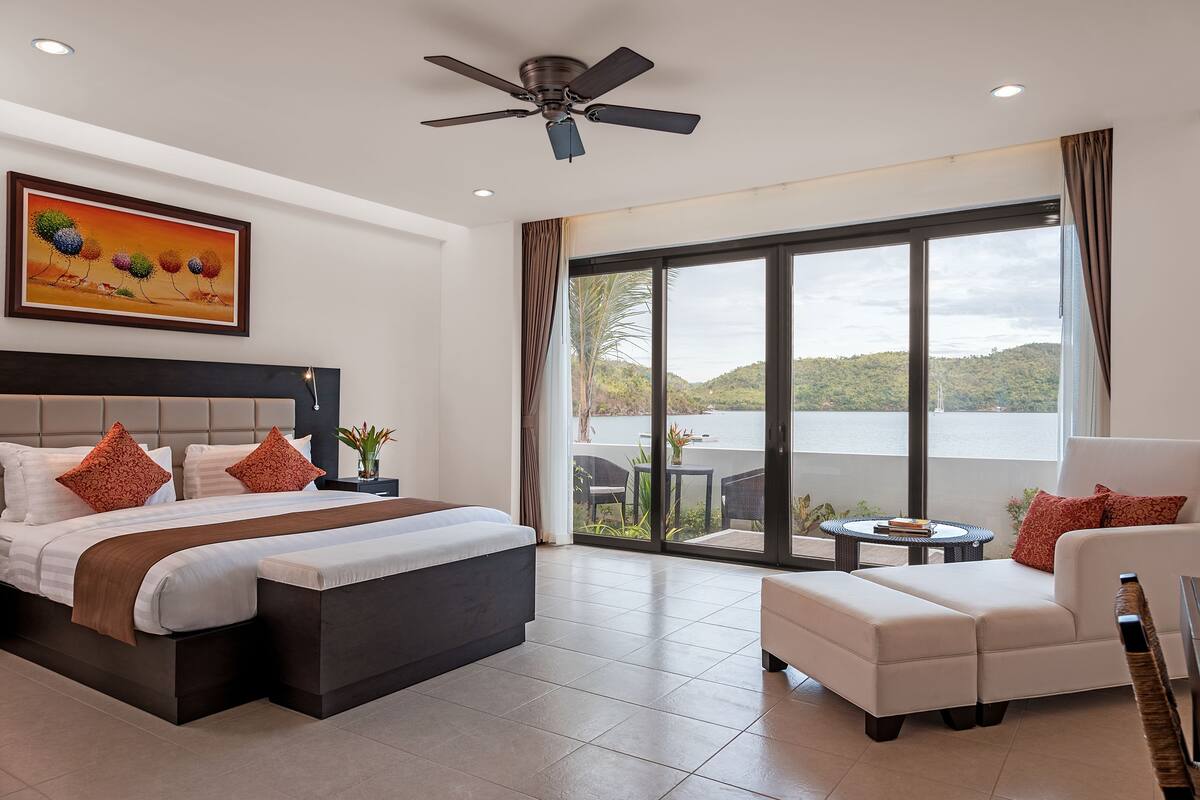 A modern hotel room with a large bed, a sofa, a ceiling fan, and a view of a lake.