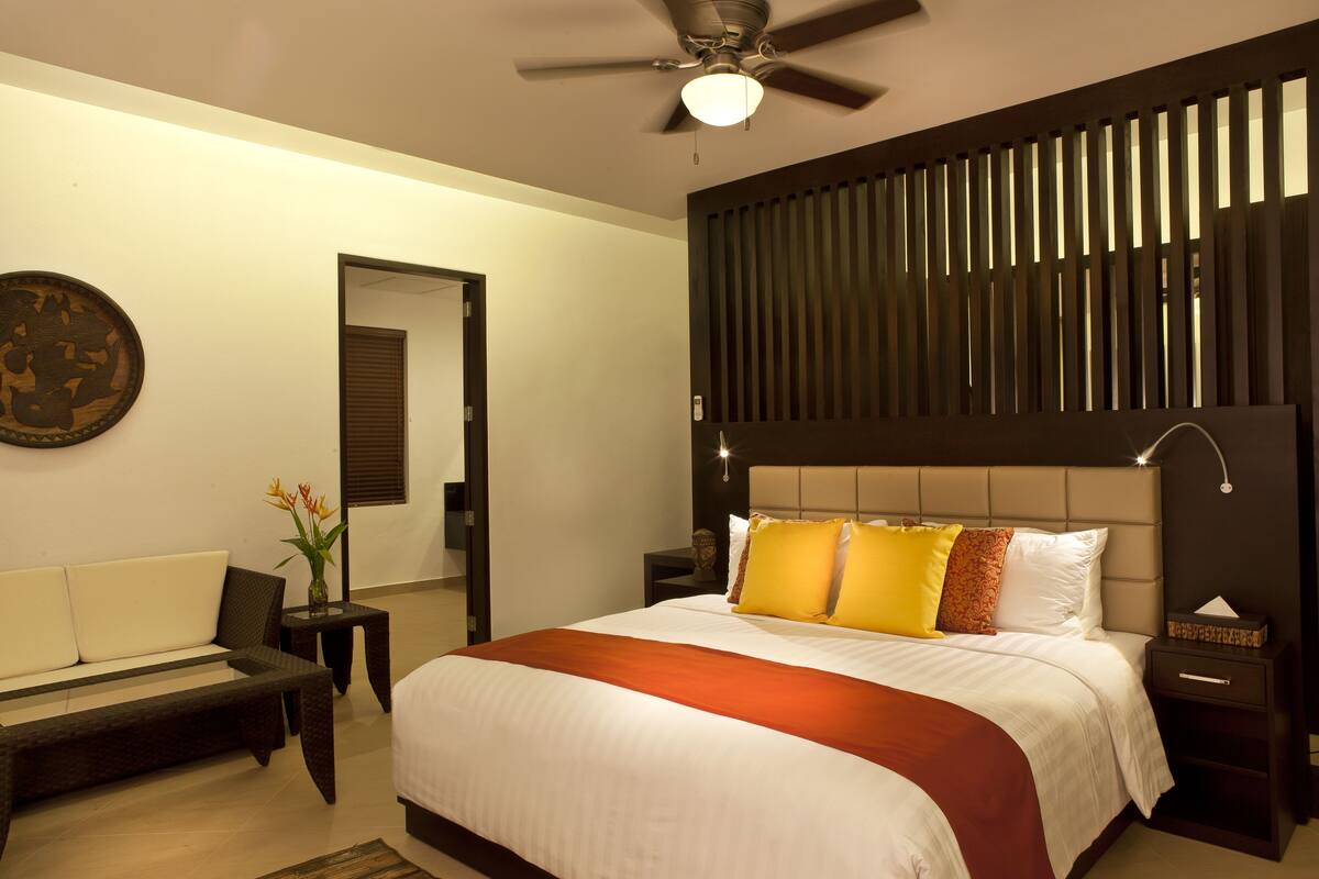 A modern hotel room with a large bed, a ceiling fan, a nightstand, a bedside table, and a sofa.