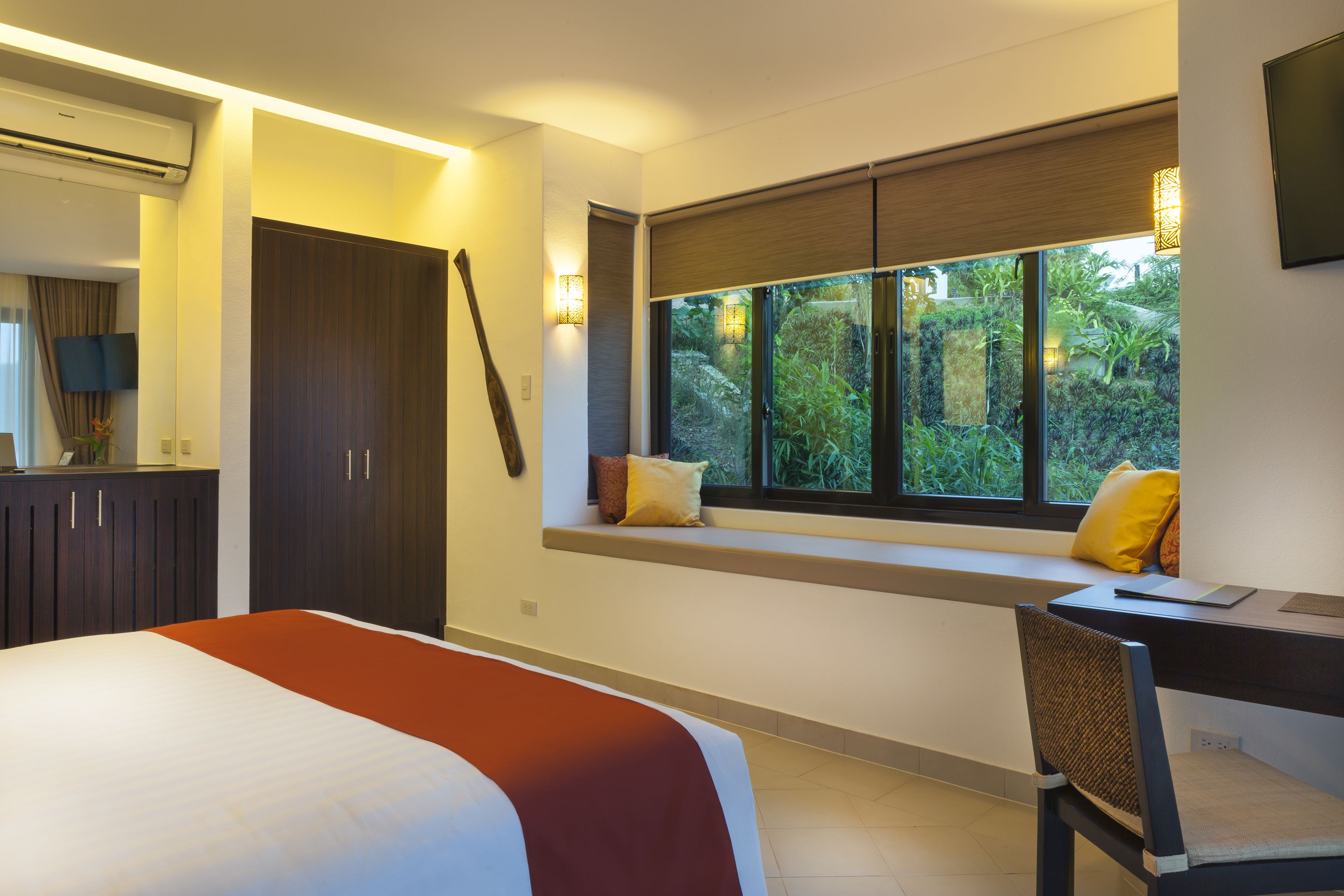 A bedroom with a bed, a desk with a chair, a window with a view of greenery, a wardrobe, and a mounted TV.