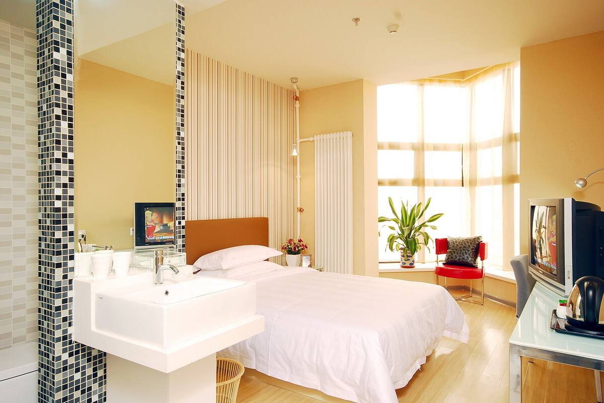 A modern bedroom with a large bed, a bedside table, a white sink, and a window with curtains.