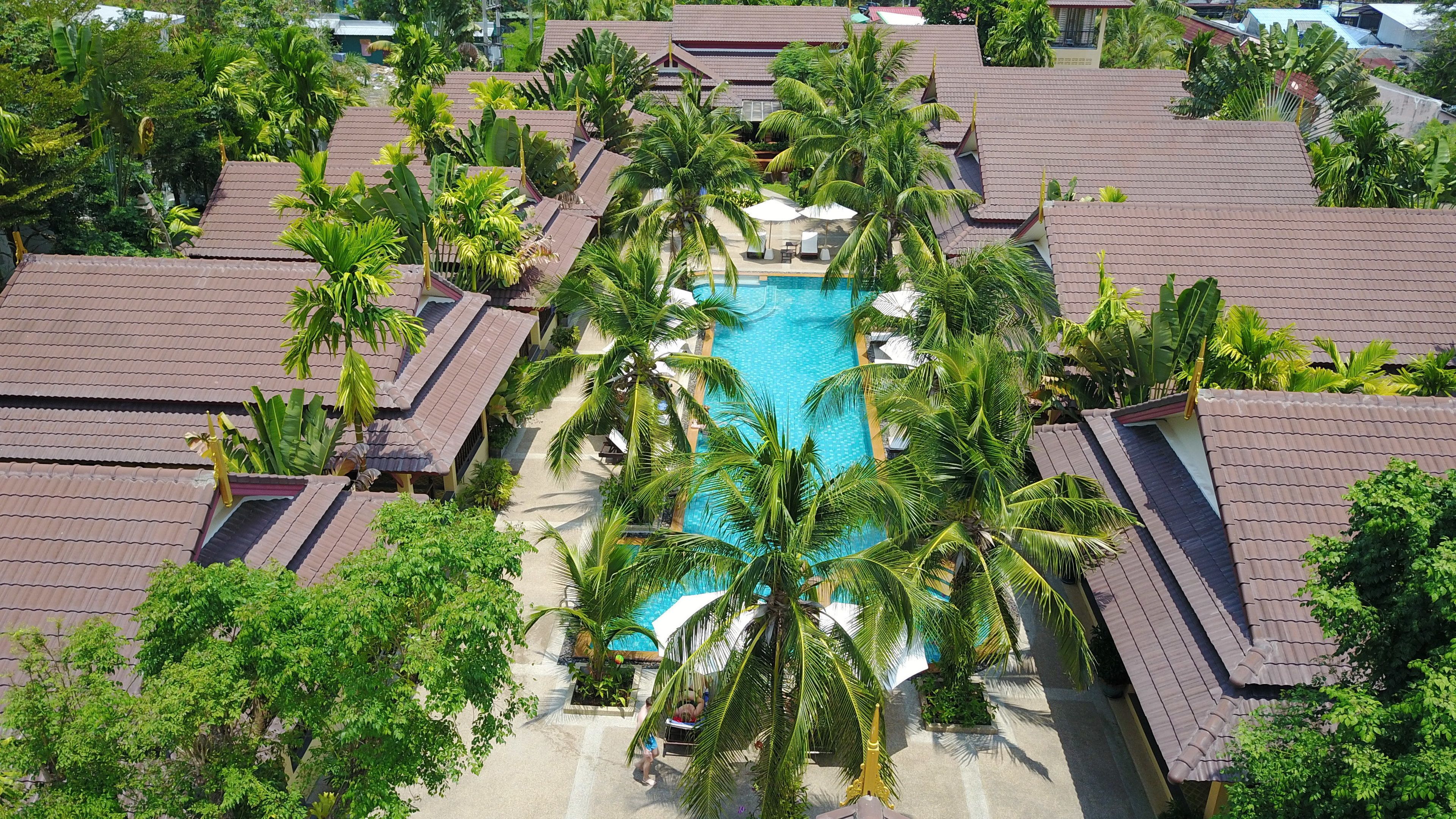 Vista aérea de un complejo turístico con varias edificaciones, una piscina central rodeada de palmeras y un cielo azul despejado.