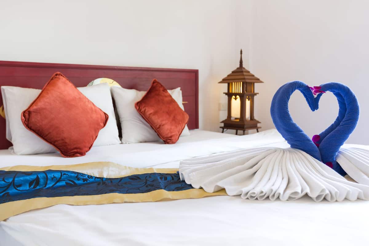 Una cama bien hecha con una toalla en forma de corazón, un cabecero de madera y un farol encendido a un lado.