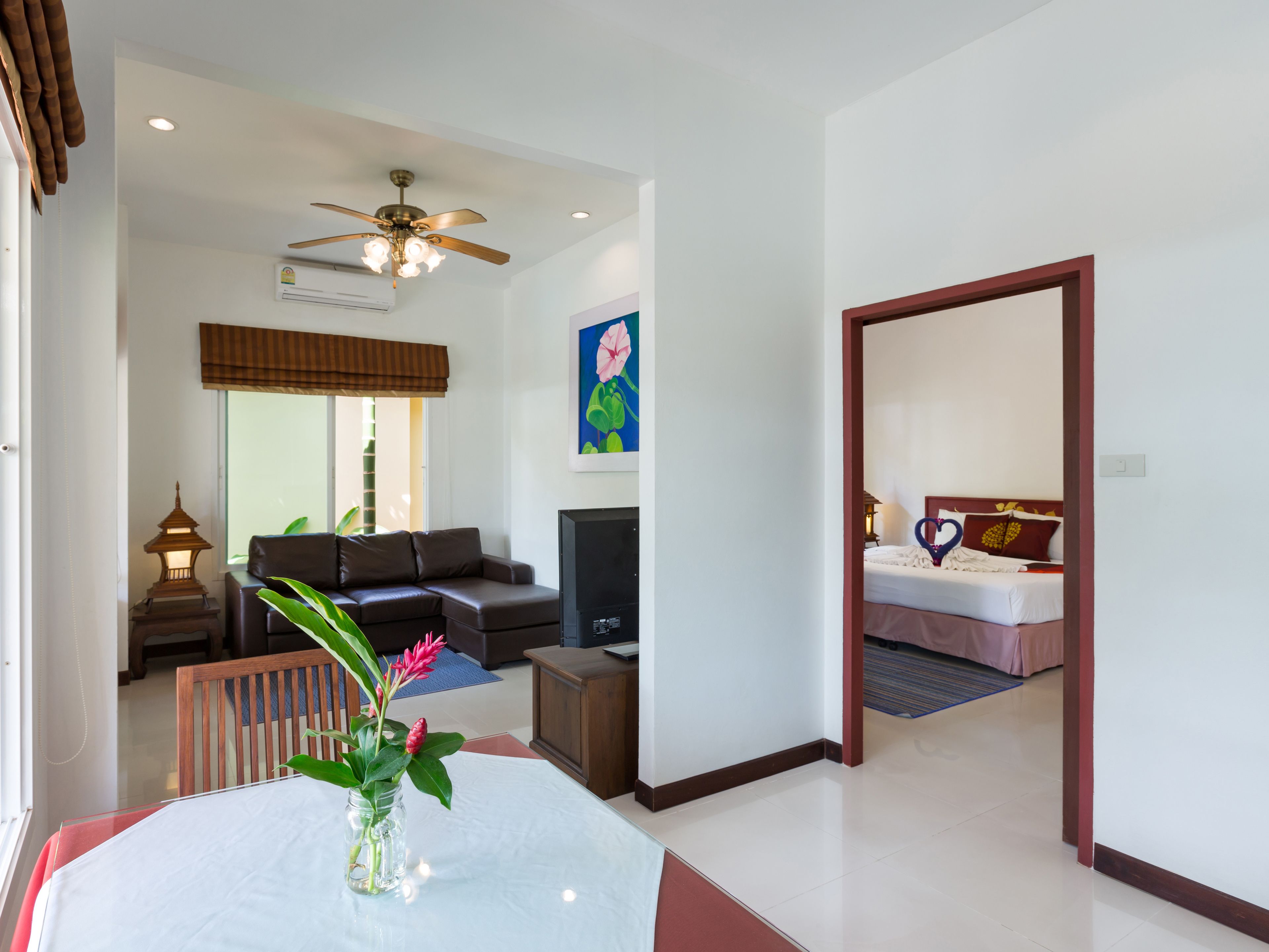 A hotel room with a bed, a TV, a ceiling fan, a dining table with a vase of flowers, and a living area with a sofa and a lamp.