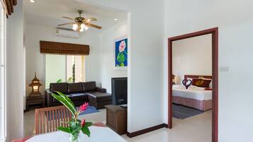 A hotel room with a bed, a TV, a ceiling fan, a dining table with a vase of flowers, and a living area with a sofa and a lamp.