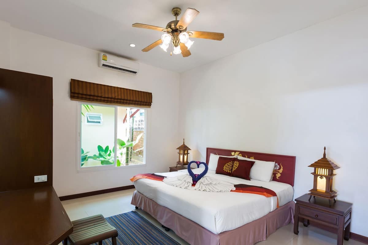 Habitación de hotel con cama, ventilador de techo, unidad de aire acondicionado, ventana con vistas, mesita de noche con farol y un pequeño banco de madera.