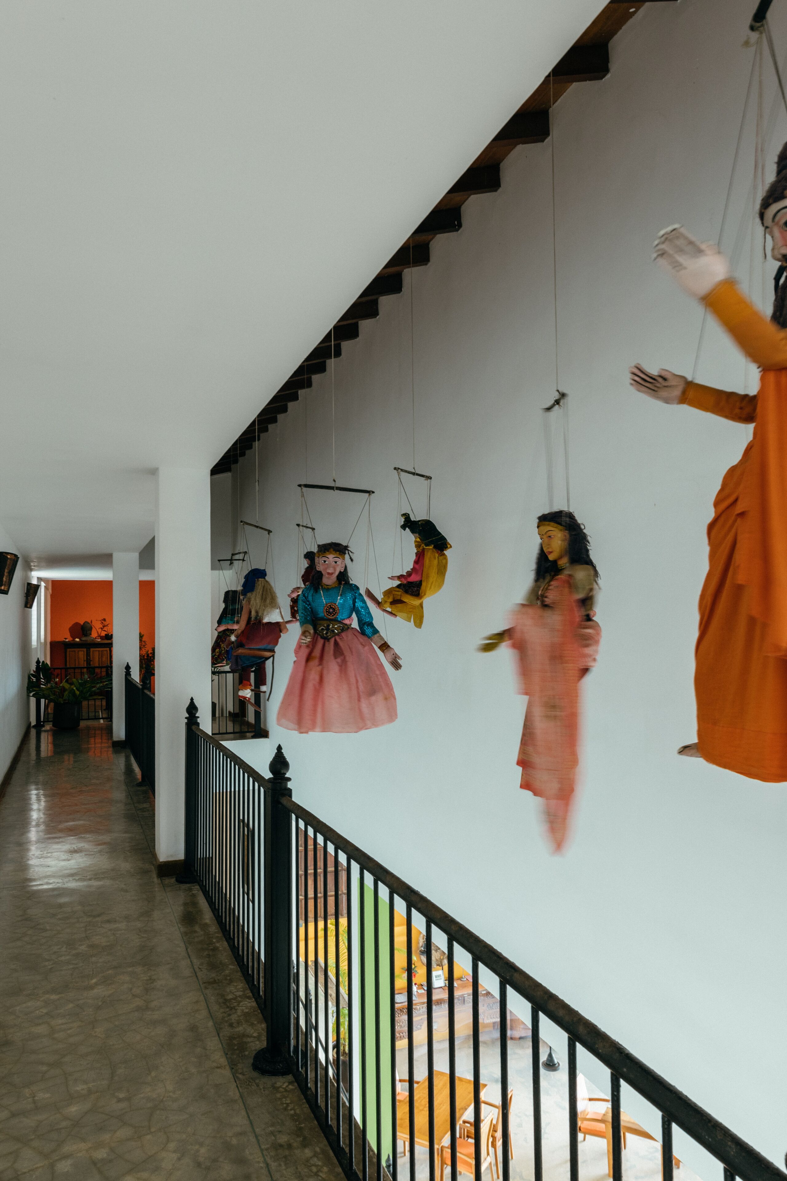 A corridor with a black railing, a white wall, and a hanging puppet display.
