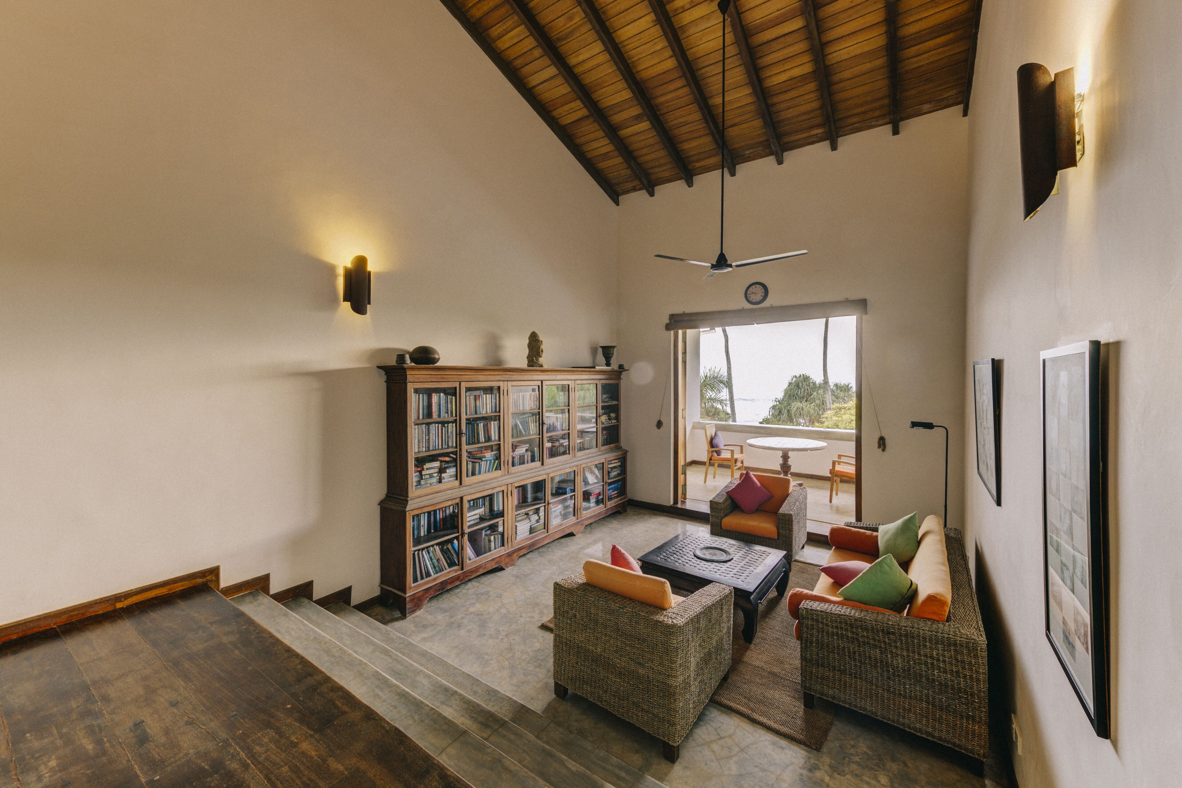 A spacious living area with a high ceiling, a large bookshelf, comfortable seating, and a view through a glass door.