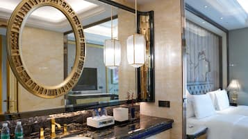 Un baño de hotel con un espejo grande, un lavabo y tocador. En la habitación contigua, hay una cama con ropa blanca.
