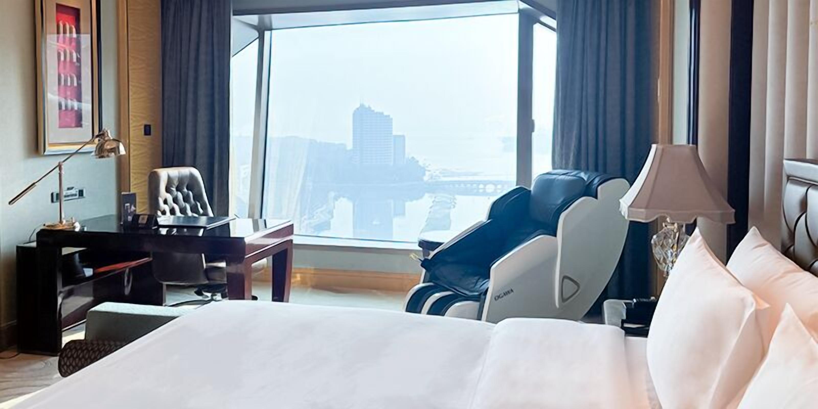 A hotel room with a bed, desk, chair, massage chair, and a view of a cityscape.