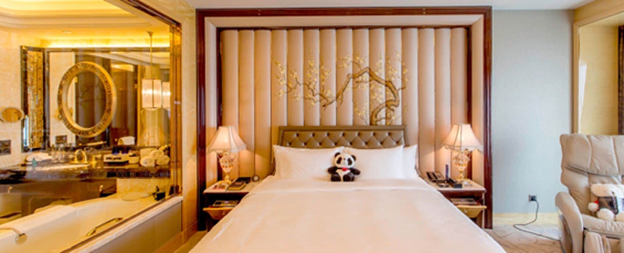 A hotel room with a large bed, a headboard with vertical panels, a nightstand with a lamp, a chair, and a mirror.