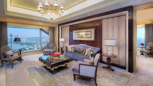 Ein luxuriöses Hotelzimmer mit großem Fenster und Blick auf die Stadt, einer bequemen Couch, eleganten Sesseln, einem Marmortisch und einem Chandelier.