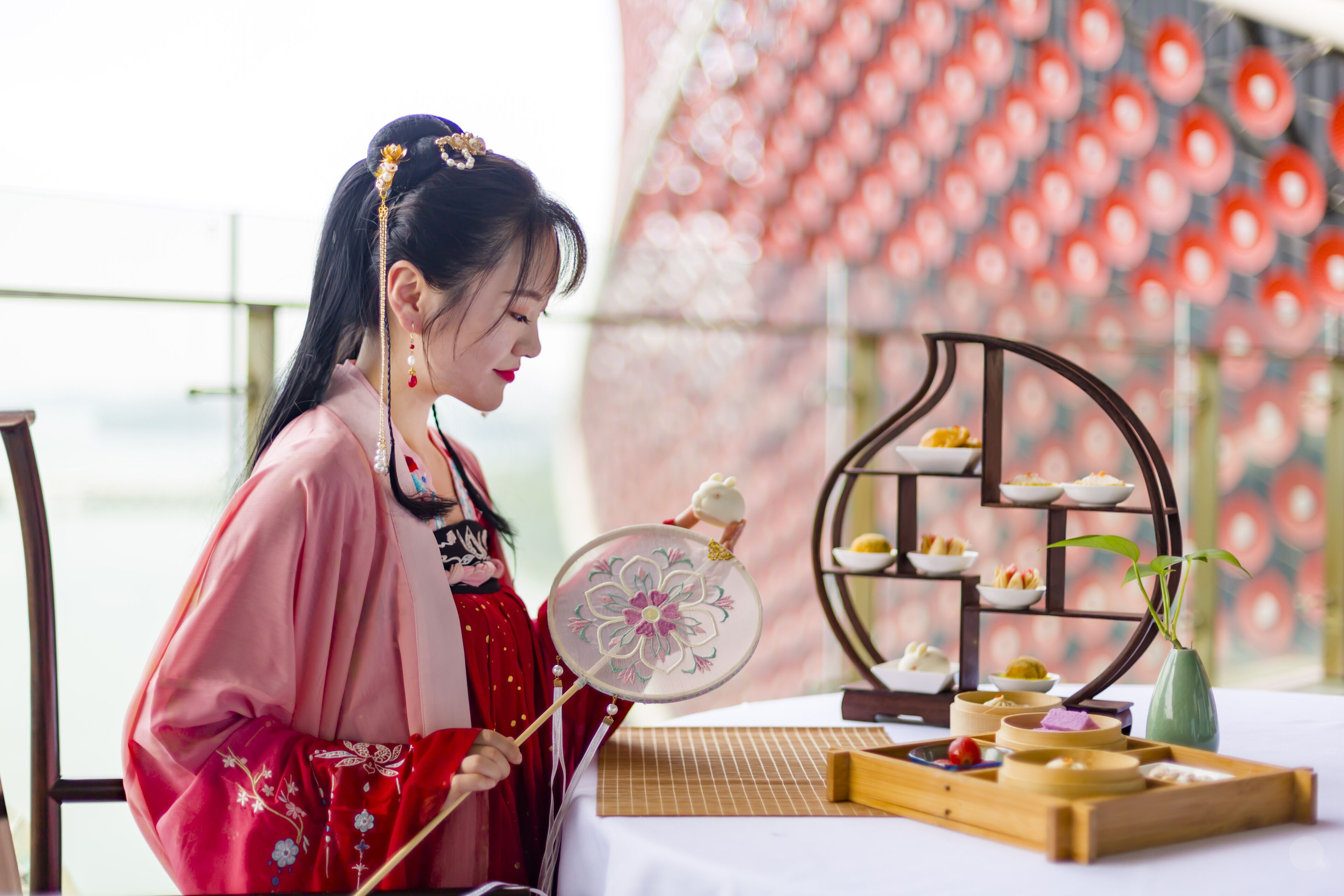 A person in traditional attire holding a decorative fan, seated at a table with a tray of food and a vase of flowers.
