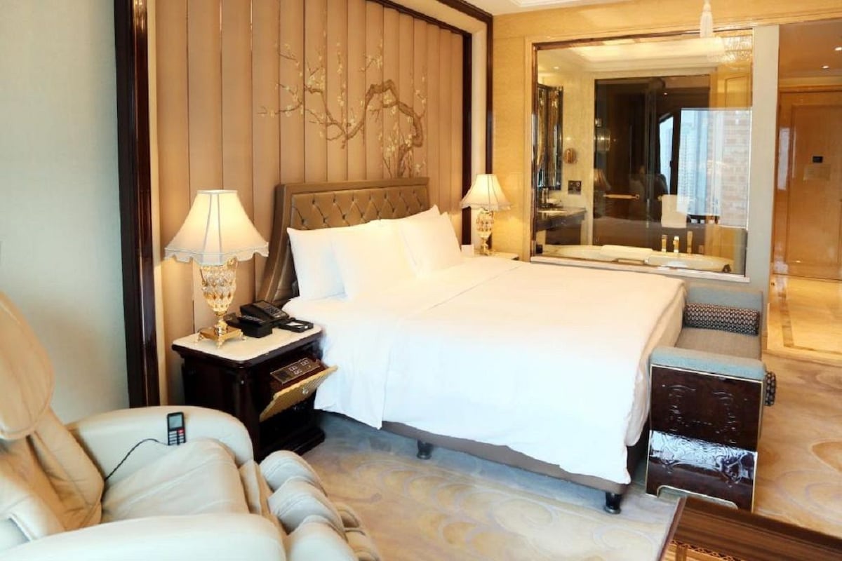 A hotel room with a large bed, two bedside tables with lamps, a mirror, a seating area with a chair, and a small wooden table.
