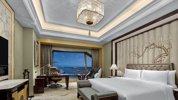 Ein Hotelzimmer mit einem großen Bett, einem Schreibtisch, einem Sessel, einer Couch, einem Fernseher und Blick auf die Stadt.