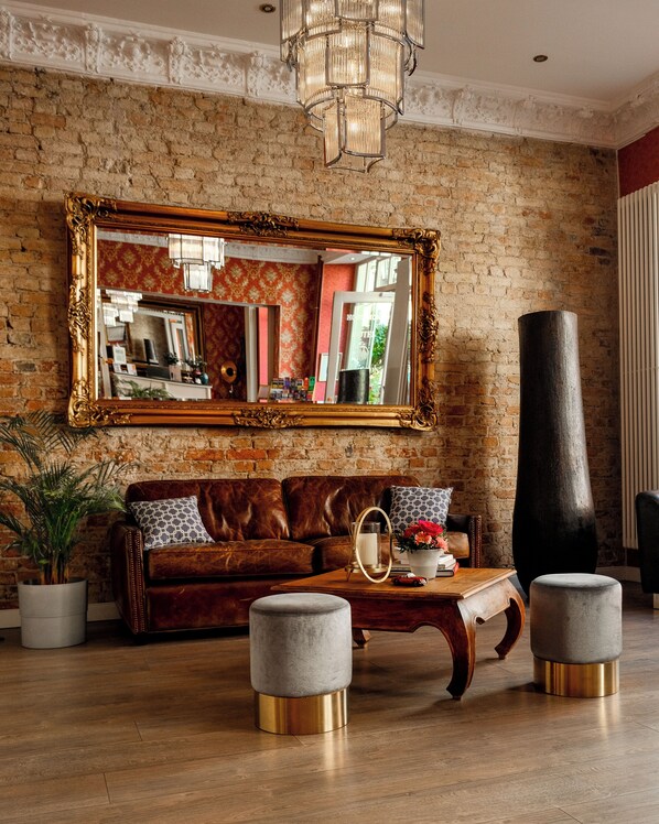 A room with a brick wall, a large mirror, a brown leather sofa, a wooden coffee table, and a tall black vase.