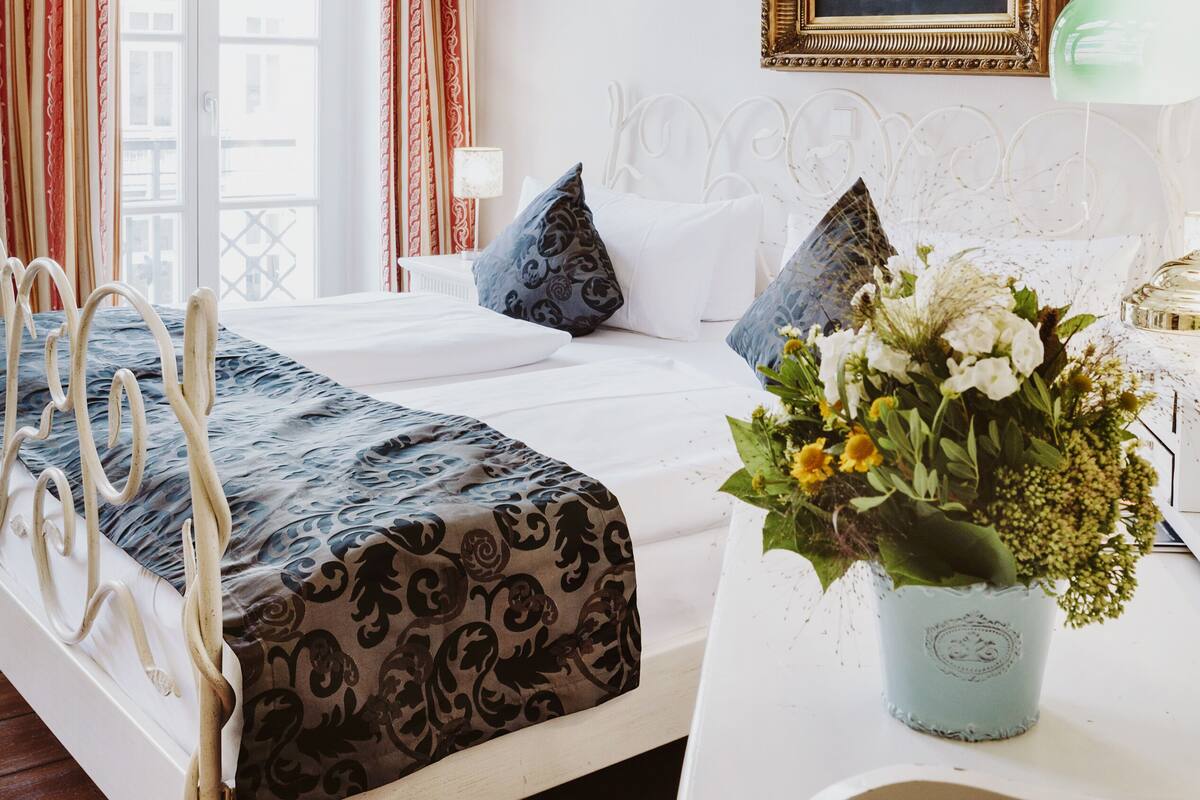 A hotel room with a bed, a window with curtains, a painting on the wall, and a vase with flowers on a table.