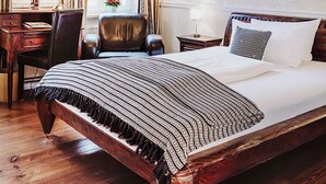 Quarto tradicional com cama grande, piso de madeira, escrivaninha, cadeira, luminária de mesa, pintura e janela com cortinas.