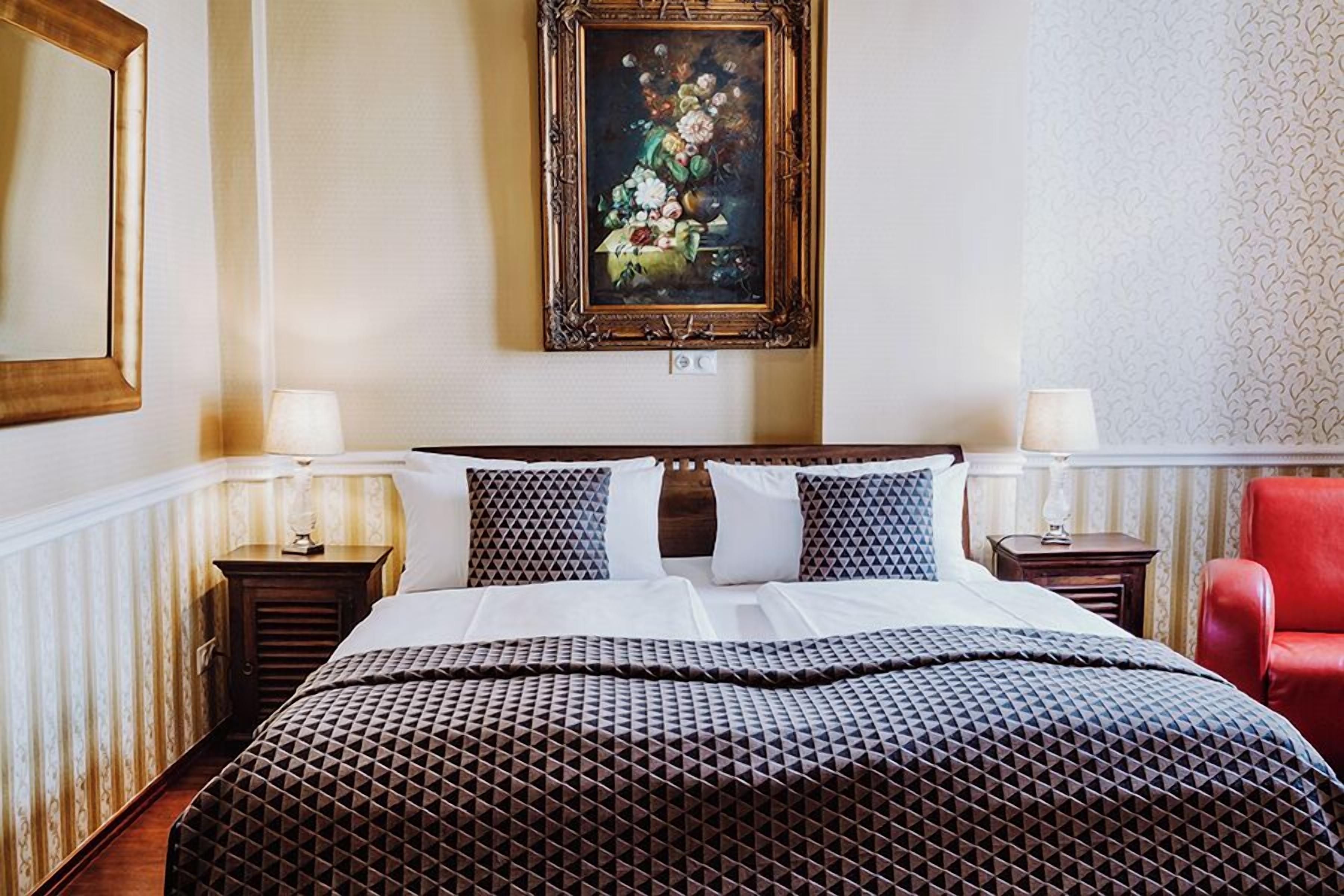 A hotel room with a large bed, two bedside tables, a red chair, and a framed painting on the wall.