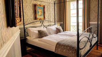 A four-poster bed with white linens and a geometric-patterned throw, set against a wallpapered wall with a framed painting.