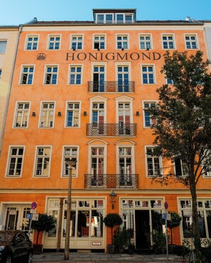 A multi-story building with the name 'HONIGMOND' on the facade, featuring balconies with metal railings, multiple windows with white frames, and a ground-level entrance with a door and potted plants.