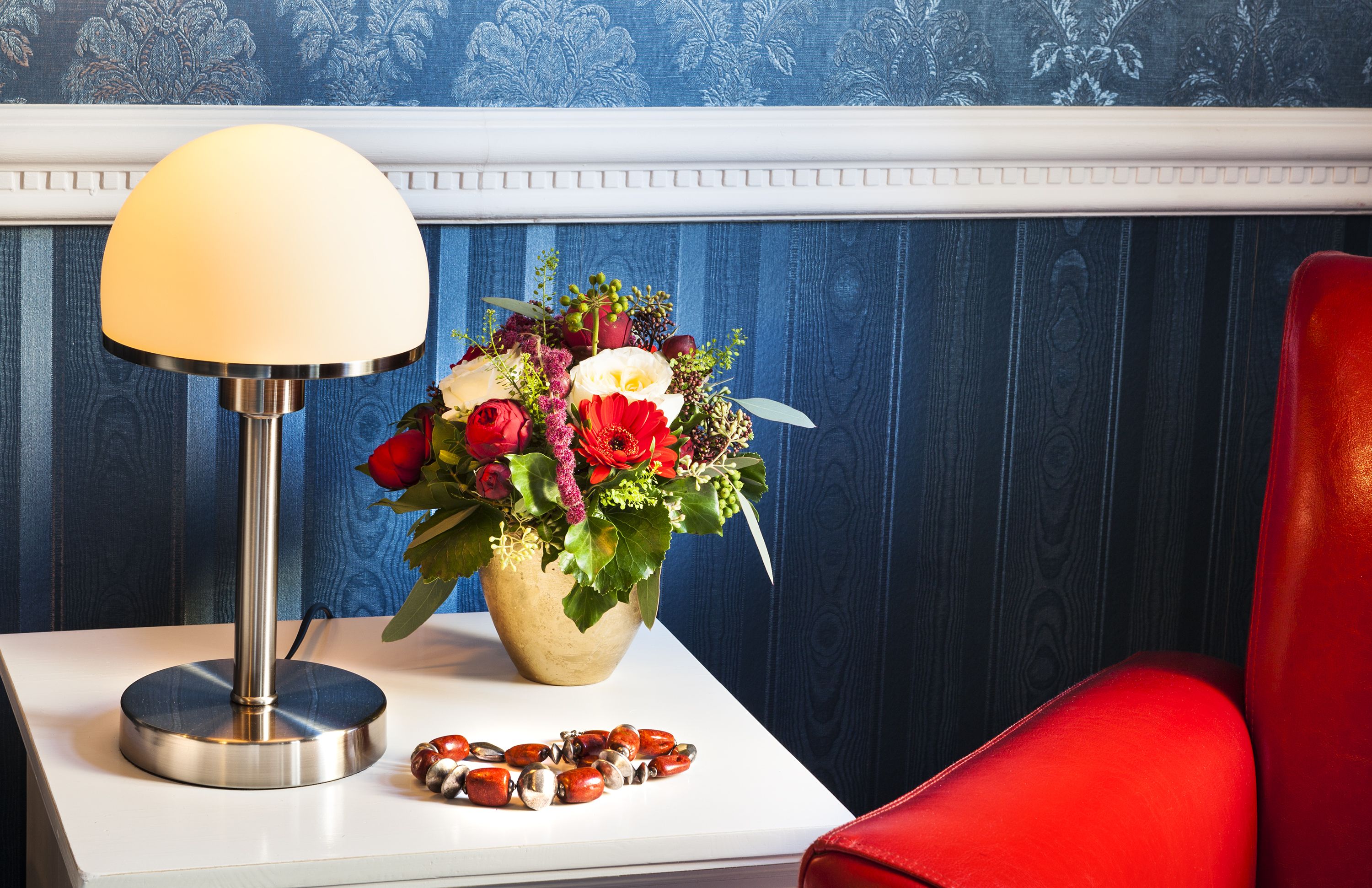 A room with a white table, a red chair, a lamp, and a floral arrangement.