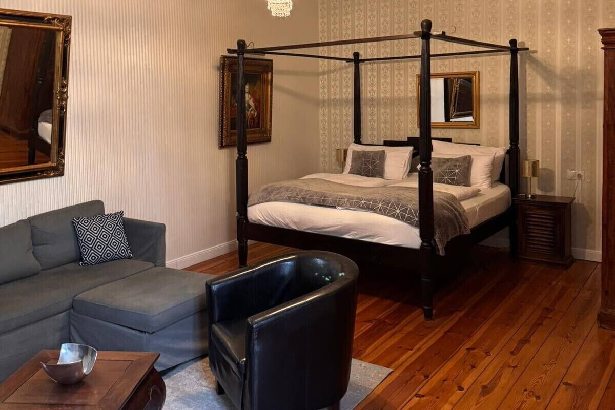 A four-poster bed, a sofa, a chair, a small table, and a mirror in a room with wooden flooring and patterned wallpaper.