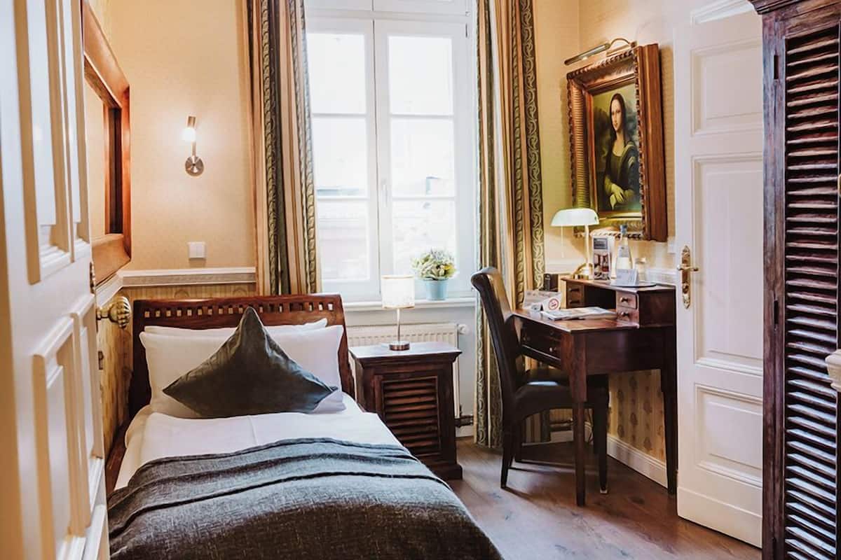 A hotel room with a bed, desk, chair, and a painting on the wall.