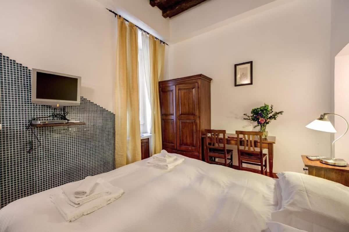 A hotel room with a bed, a desk with a chair, a wooden wardrobe, a TV mounted on the wall, and a window with curtains.