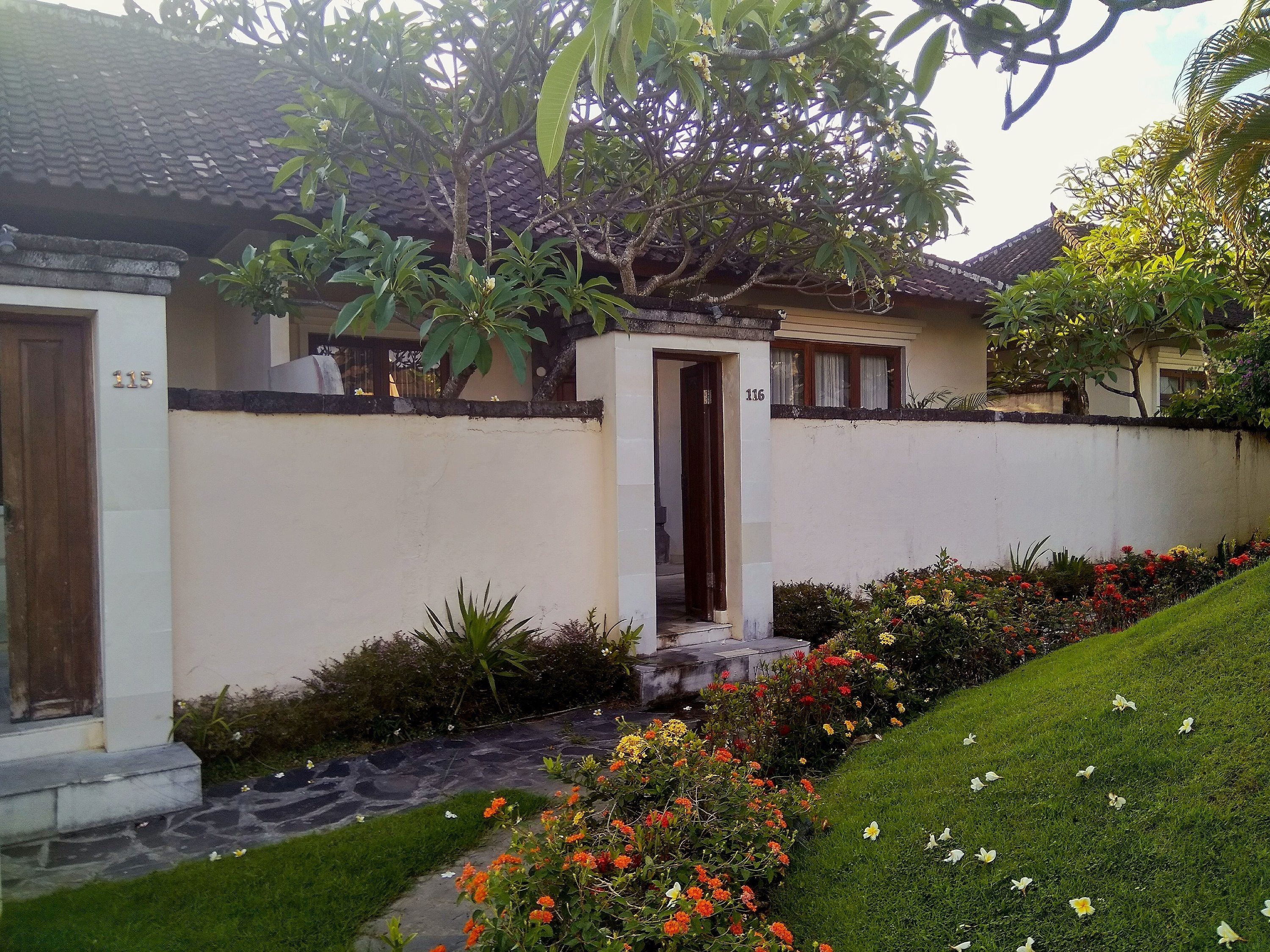 Une maison avec un toit en tuiles, un mur blanc et un petit jardin avec des fleurs et de l’herbe.