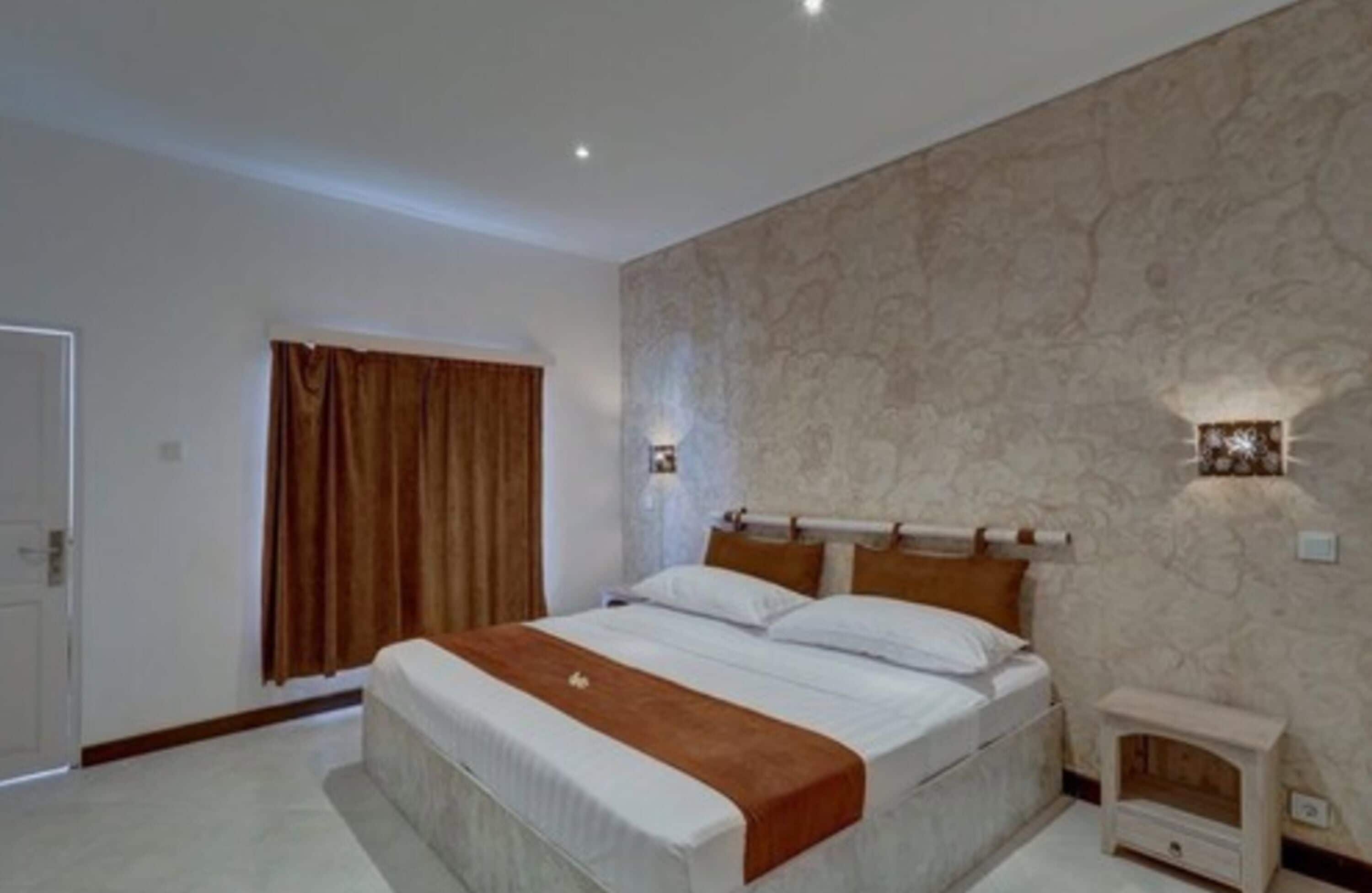 A bedroom with a large bed, a nightstand, a wall-mounted lamp, and a window with curtains.