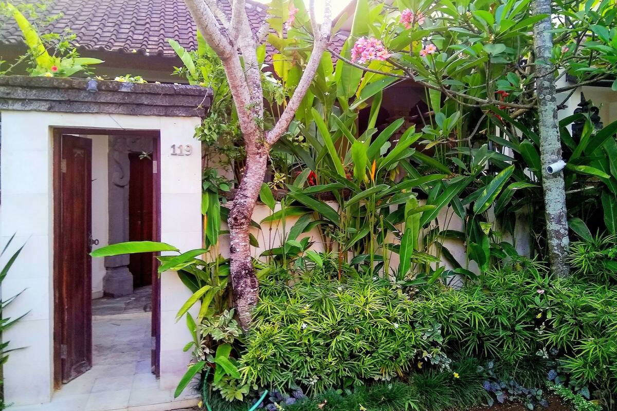 A traditional house with a tiled roof, a door labeled '119', and lush greenery including various plants and a tree with pink flowers.