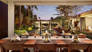 Uma área de refeições com vista para uma piscina e jardim, com uma mesa posta para quatro pessoas e paisagem tropical.