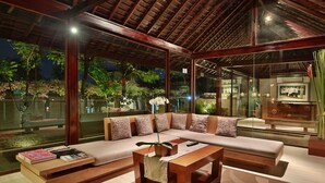 Uma sala de estar moderna com um sofá grande, uma mesa de centro e uma parede de vidro que oferece vista para um jardim.