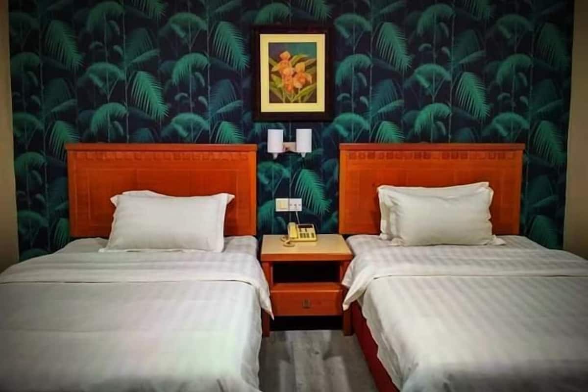 Ein Hotelzimmer mit zwei Betten, jeweils mit weißen Bettwäsche und passenden Kissen. Zwischen den Betten befindet sich ein Nachttisch mit einer Lampe und einem Telefon. Die Wände sind mit einem tropischen Blattmuster verziert.