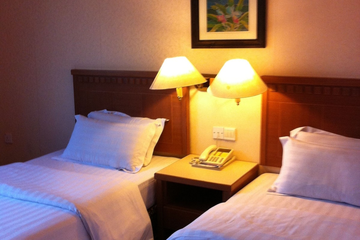 Ein Hotelzimmer mit zwei Betten, jeweils mit weißen Laken und Kissen. Zwei Nachttischlampen, ein an der Wand befestigtes Telefon und ein gerahmtes Bild über den Lampen sind vorhanden.