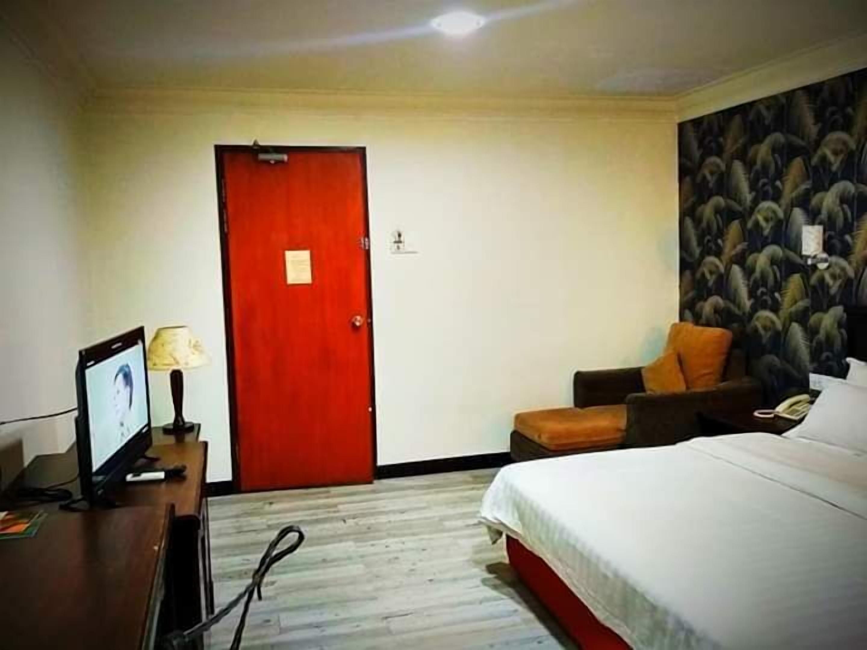 A hotel room with a bed, a desk with a computer, a chair, and a red door.
