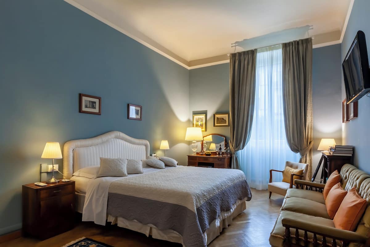 Habitación de hotel con cama, mesitas de noche, una silla, sofá, televisión y ventana con cortinas.