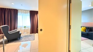 Ein modernes Hotelzimmer mit einem großen Fenster, einer Sitzecke mit einem Sessel und einem kleinen Tisch sowie einem separaten Wohnbereich mit einer Couch und Kissen.